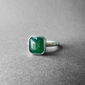 K&ouml;nnte beinhalten: Ein silberfarbener Ring mit einem gro&szlig;en, rechteckigen, smaragdgeschliffenen gr&uuml;nen Edelstein in einer glatten Zargenfassung. Die Ringschiene ist poliert und metallisch und zeigt ein klares, schlichtes Design, das die leuchtend gr&uuml;ne Farbe betont.