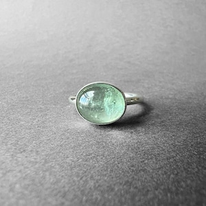 Op de afbeelding: Een zilveren ring met een grote, ovale, lichtgroene edelsteen. De edelsteen is gezet in een gepolijste zilveren band. De ring wordt getoond op een grijze ondergrond, wat het elegante ontwerp en de kleur benadrukt.