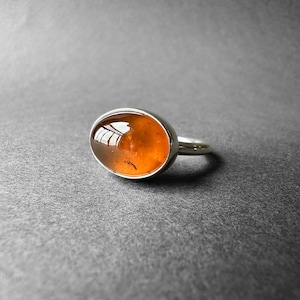 May include: A silver ring with an oval-shaped amber gemstone. The gemstone is a deep orange color and has a slight shimmer.