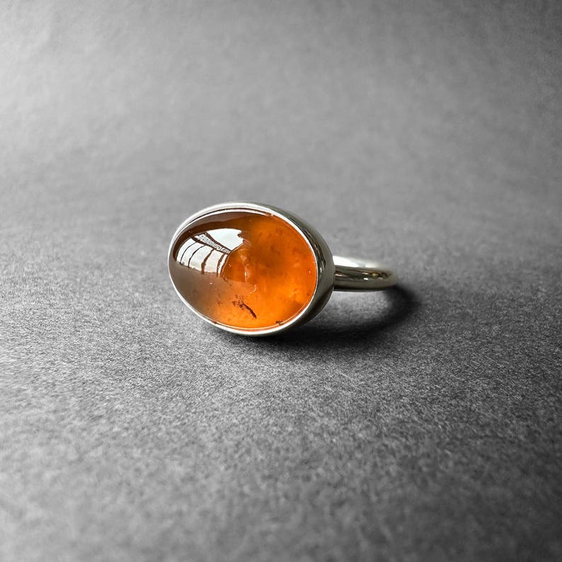 May include: A silver ring with an oval-shaped amber gemstone. The gemstone is a deep orange color and has a slight shimmer.