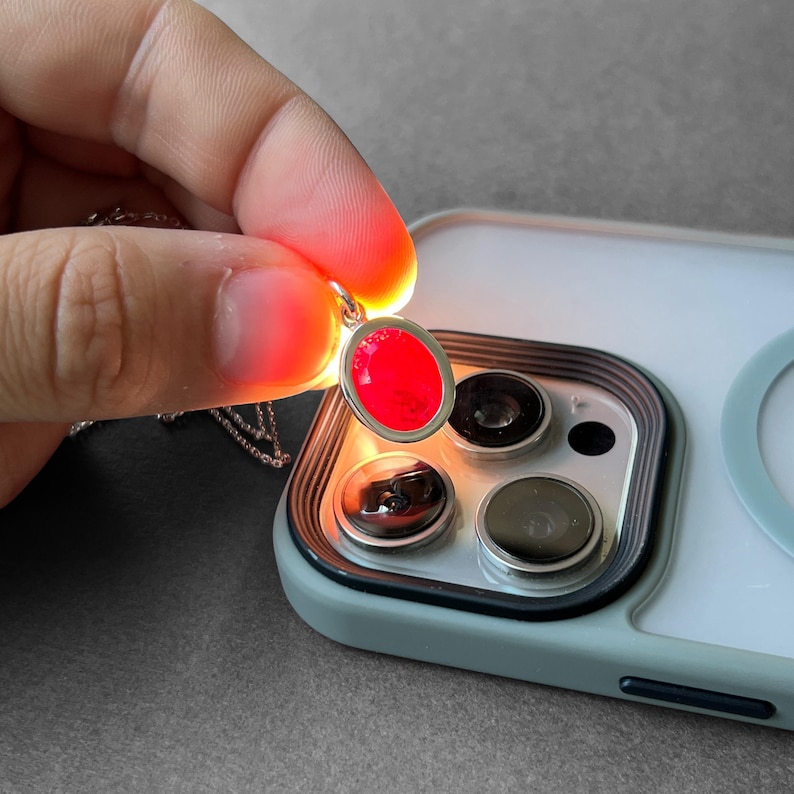 Op de afbeelding: Een zilveren hanger met een ovale, doorschijnende rode steen die boven de cameralens van een smartphone wordt gehouden. Het licht van de flitser van de telefoon schijnt door de rode steen, wat een helder, gloeiend effect in de hanger cre&euml;ert.