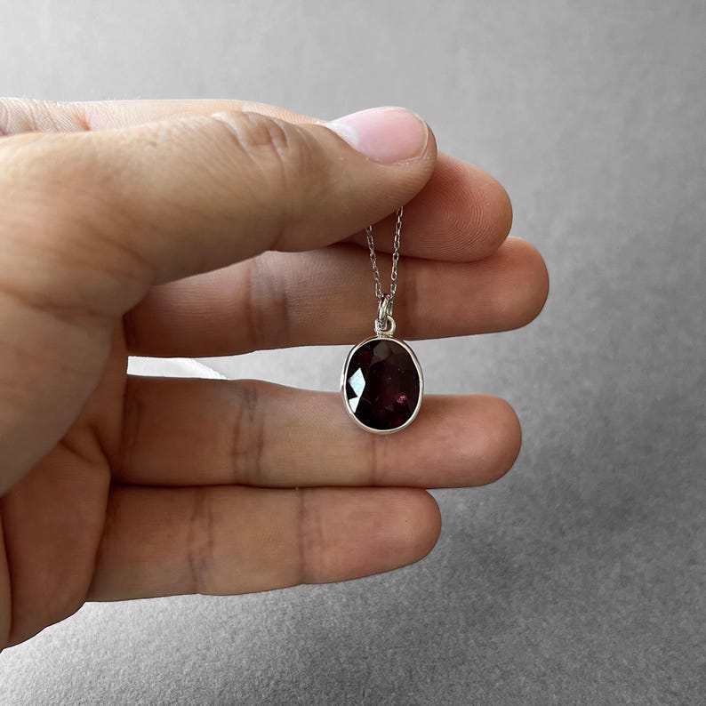 Op de afbeelding: Een zilveren ketting met een ovaalvormige, gefacetteerde donkerrode edelsteen hanger. De steen is gezet in een eenvoudige zilveren rand en hangt aan een fijne zilveren ketting, met een diepe bordeauxrode kleur en reflecterende vlakken.