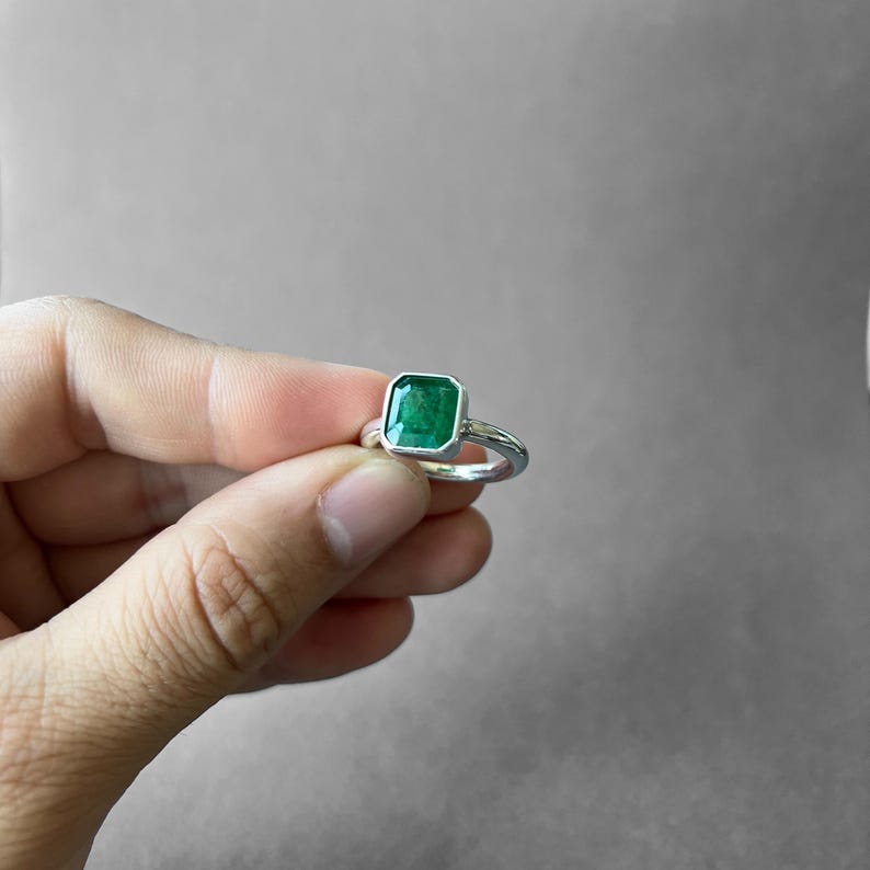 K&ouml;nnte beinhalten: Ein Silberring mit einem rechteckigen, smaragdgeschliffenen gr&uuml;nen Edelstein in einer glatten Zargenfassung. Die Ringschiene besteht aus poliertem Silber und h&auml;lt den leuchtend gr&uuml;nen Stein sicher in der Mitte, was ein schlichtes Design ergibt.