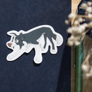May include: A die-cut sticker featuring a cartoon illustration of a black and white border collie dog in a playful, crouching stance. The dog has large eyes, a brown nose, and white markings on its chest, legs, and tail tip, set against a dark background.