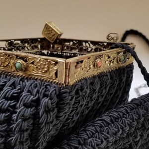 Puede incluir: Un bolso de noche de crochet negro con un marco de metal dorado y piedras de colores. El bolso tiene una correa de cadena y un peque&ntilde;o cierre cuadrado.