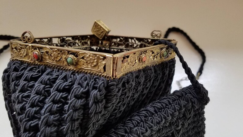 Puede incluir: Un bolso de noche de crochet negro con un marco de metal dorado y piedras de colores. El bolso tiene una correa de cadena y un peque&ntilde;o cierre cuadrado.