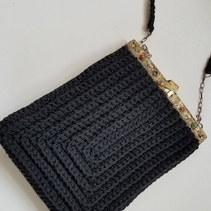 Puede incluir: Un bolso de noche de crochet negro con una correa de cadena dorada y un cierre decorativo con piedras de colores.