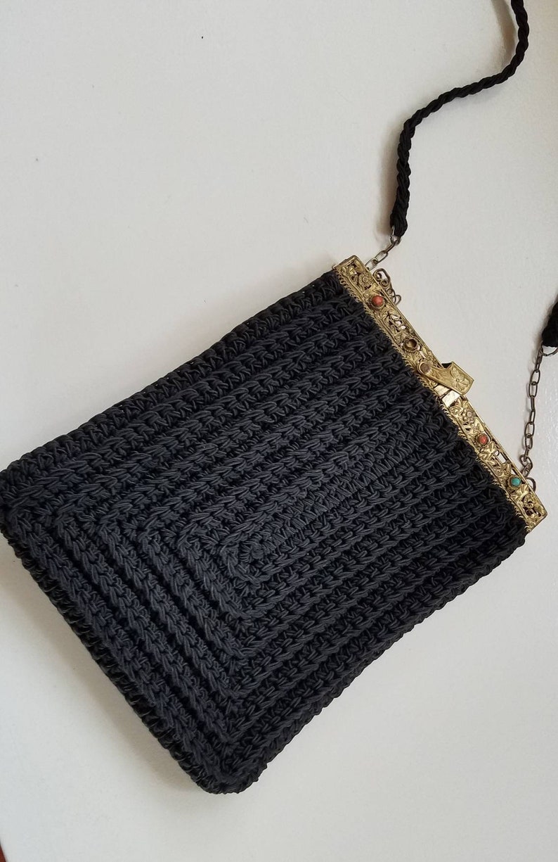 Puede incluir: Un bolso de noche de crochet negro con una correa de cadena dorada y un cierre decorativo con piedras de colores.