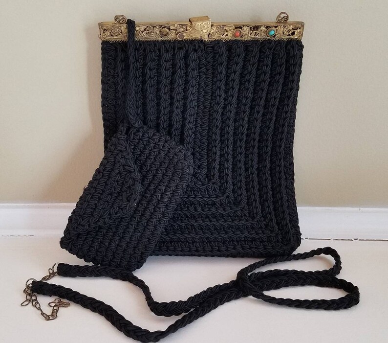 Puede incluir: Un bolso de crochet negro con un cierre de metal dorado y una correa de crochet negra a juego. El bolso tiene un peque&ntilde;o bolso de crochet a juego adjunto.