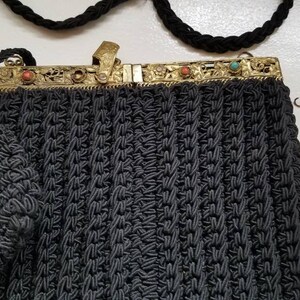 Puede incluir: Un bolso de noche de crochet negro con un cierre de metal dorado y piedras de colores. El bolso tiene una correa de cadena.