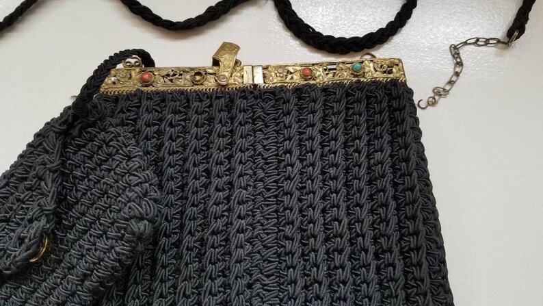 Puede incluir: Un bolso de noche de crochet negro con un cierre de metal dorado y piedras de colores. El bolso tiene una correa de cadena.