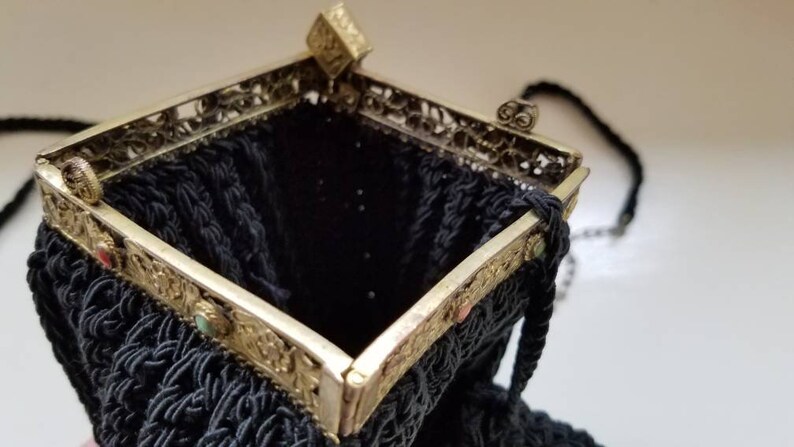 Puede incluir: Un bolso de noche negro de cuentas con un marco de metal plateado y detalles ornamentados. El bolso tiene una correa de cadena y un cierre de broche.