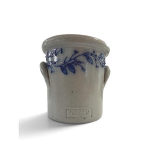 May include: A beige stoneware crock featuring two side handles and a blue hand-painted vine pattern around the upper rim. A rectangular stamp on the front reads Salmon Falls Stoneware 1995 in raised lettering on the ceramic surface.