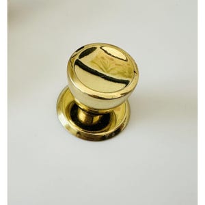 May include: A gold-toned door knob with a round, smooth top and a slightly raised rim. The knob is attached to a round base.