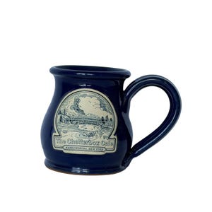 May include: A navy blue ceramic mug with a curved handle. The mug features a white and black illustration of a bridge and landscape, with the text "The Chatterbox Cafe, Narrowsburg, New York."
