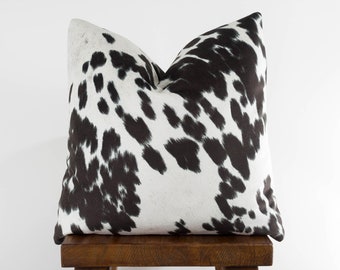 May include: A black and white cowhide patterned throw pillow sits on a wooden stool. The pillow is a decorative accent for a home.