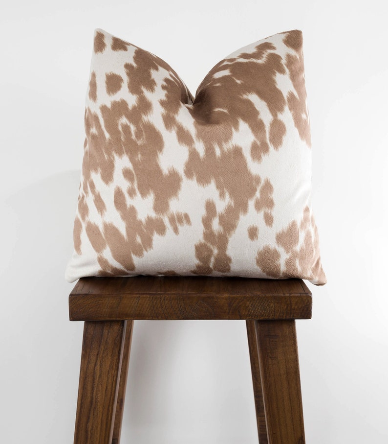 May include: A brown and white cowhide patterned throw pillow sits on a wooden stool. The pillow is a soft, plush fabric with a subtle sheen.