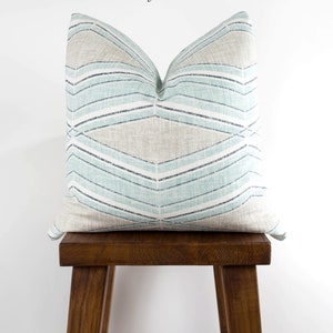 Puede incluir: Una almohada de tiro azul claro y beige con un patr&oacute;n geom&eacute;trico. La almohada est&aacute; sentada en un taburete de madera. El texto "Biscayne Harbor" est&aacute; escrito sobre la almohada.