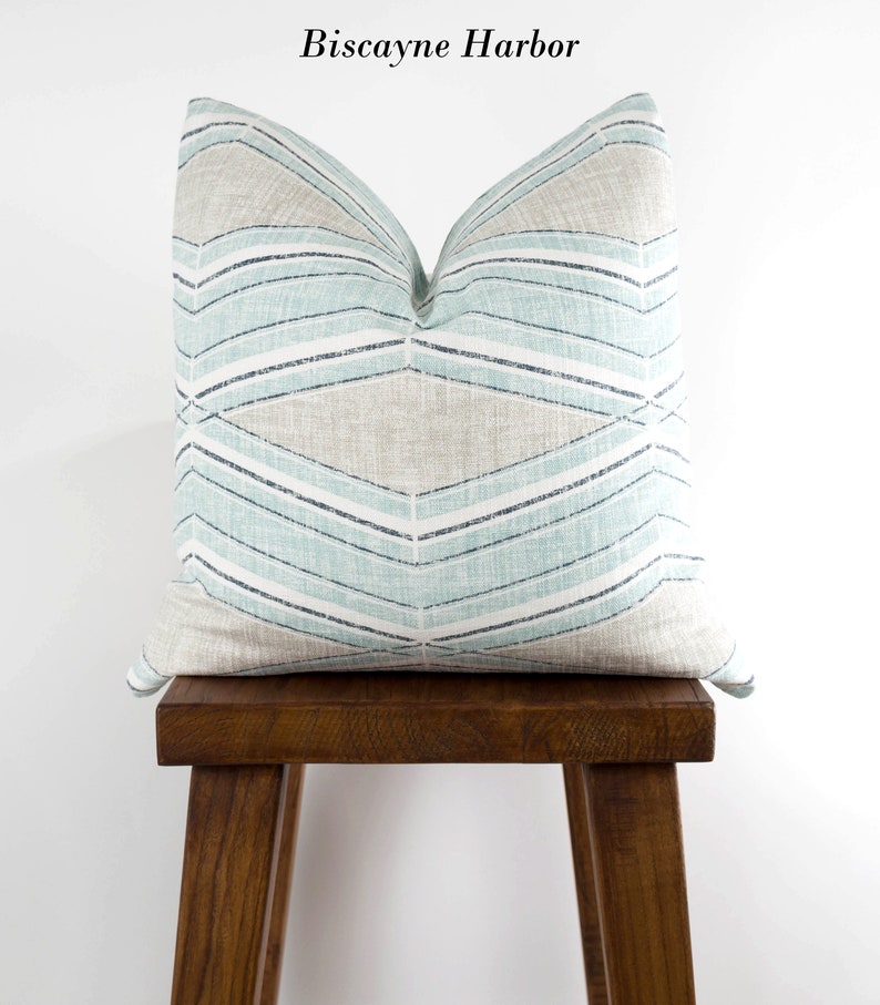 Puede incluir: Una almohada de tiro azul claro y beige con un patr&oacute;n geom&eacute;trico. La almohada est&aacute; sentada en un taburete de madera. El texto "Biscayne Harbor" est&aacute; escrito sobre la almohada.