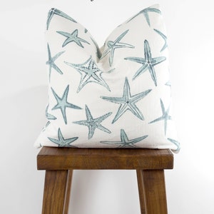 Puede incluir: Una almohada blanca con un patr&oacute;n de estrellas de mar azules. La almohada est&aacute; sentada en un taburete de madera. El texto "Starfish Harbor" est&aacute; escrito encima de la almohada.