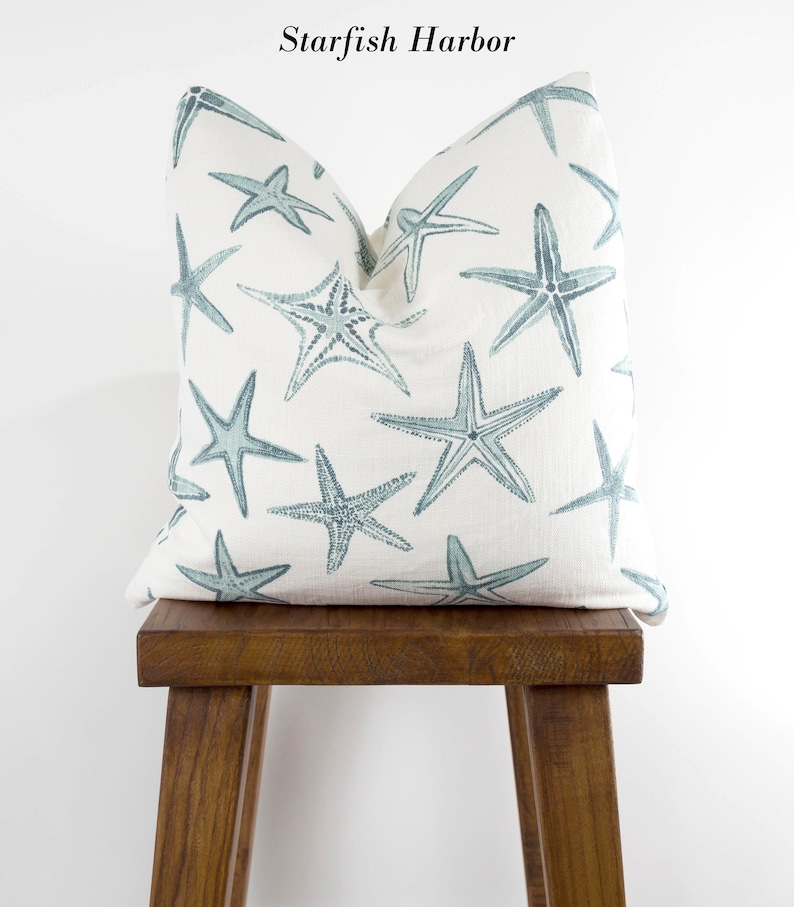Puede incluir: Una almohada blanca con un patr&oacute;n de estrellas de mar azules. La almohada est&aacute; sentada en un taburete de madera. El texto "Starfish Harbor" est&aacute; escrito encima de la almohada.
