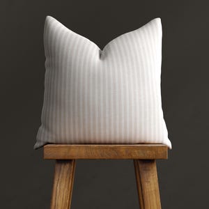 May include: A white and beige striped cushion sits on a wooden stool against a dark grey background. The cushion is made of a soft, textured fabric and has a subtle sheen. The stool is made of natural wood and has a simple, rustic design.