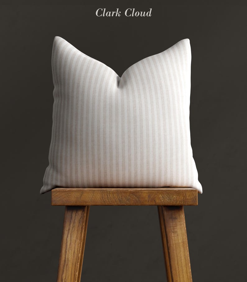 May include: A white and beige striped cushion sits on a wooden stool against a dark grey background. The cushion is made of a soft, textured fabric and has a subtle sheen. The stool is made of natural wood and has a simple, rustic design.
