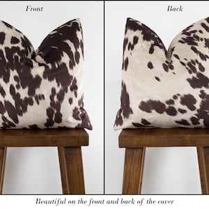 May include: A brown and white cowhide patterned pillow cover. The front and back of the cover feature the same pattern. The pillow is displayed on a wooden stool.