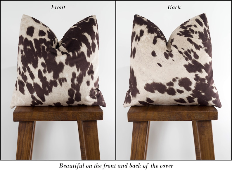 May include: A brown and white cowhide patterned pillow cover. The front and back of the cover feature the same pattern. The pillow is displayed on a wooden stool.