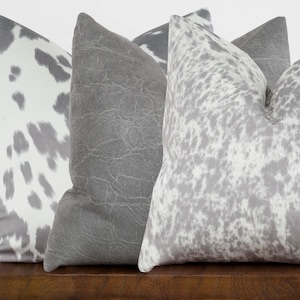 May include: Three decorative throw pillows with a cowhide pattern in shades of gray and white. The pillows are arranged on a wooden surface.