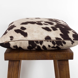 May include: A brown and white cowhide patterned throw pillow with a zipper closure. The pillow is sitting on a wooden stool.