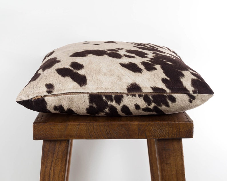 May include: A brown and white cowhide patterned throw pillow with a zipper closure. The pillow is sitting on a wooden stool.