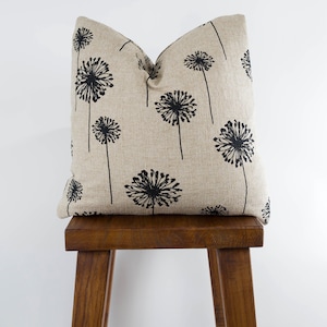May include: A beige linen pillow with a black dandelion print. The pillow is sitting on a wooden stool.