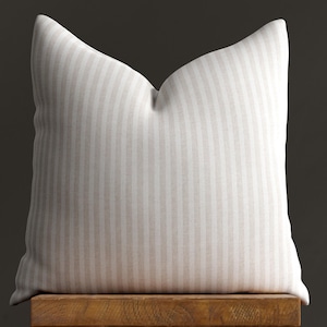 May include: A white and beige striped throw pillow with a square shape. The pillow is sitting on a wooden surface.