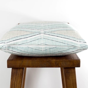 Puede incluir: Una almohada de tiro azul claro y beige con un patr&oacute;n geom&eacute;trico. La almohada tiene una cremallera y est&aacute; sentada en un taburete de madera.