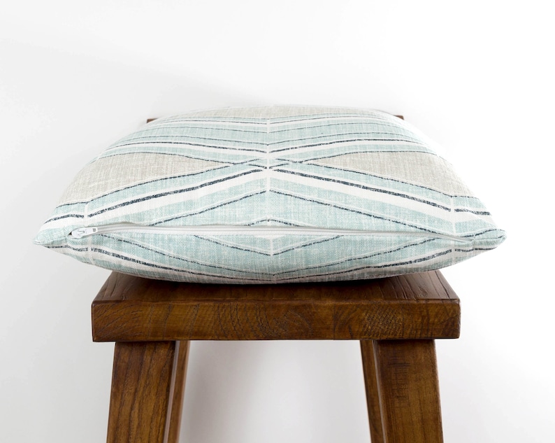 Puede incluir: Una almohada de tiro azul claro y beige con un patr&oacute;n geom&eacute;trico. La almohada tiene una cremallera y est&aacute; sentada en un taburete de madera.