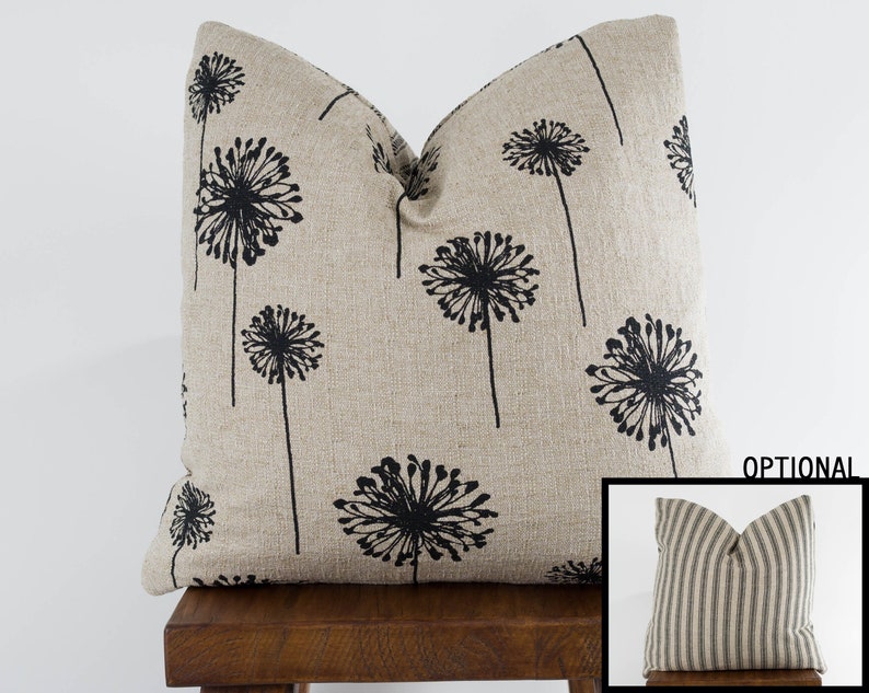 May include: A beige linen pillow with a black dandelion print. The pillow is sitting on a wooden stool.  Optional: A black and white striped pillow is shown in the bottom right corner of the image.