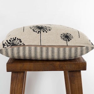May include: A beige linen pillow with a black dandelion print and a black and white striped back. The pillow is sitting on a wooden stool.
