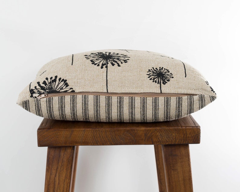 May include: A beige linen pillow with a black dandelion print and a black and white striped back. The pillow is sitting on a wooden stool.