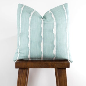 Puede incluir: Una almohada de tiro azul claro con rayas blancas abstractas. La almohada est&aacute; sentada en un taburete de madera. El texto "Canal Harbor" est&aacute; escrito sobre la almohada.