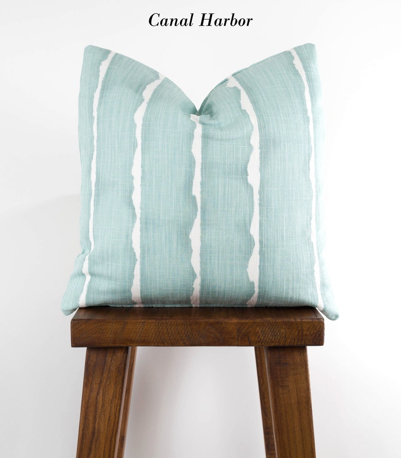 Puede incluir: Una almohada de tiro azul claro con rayas blancas abstractas. La almohada est&aacute; sentada en un taburete de madera. El texto "Canal Harbor" est&aacute; escrito sobre la almohada.