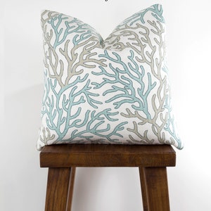 Puede incluir: Una almohada decorativa con un patr&oacute;n de arrecife de coral en tonos azules y beige. La almohada est&aacute; sentada en un taburete de madera.