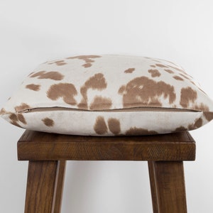 May include: A brown and white cowhide patterned throw pillow with a zipper closure. The pillow is sitting on a wooden stool.