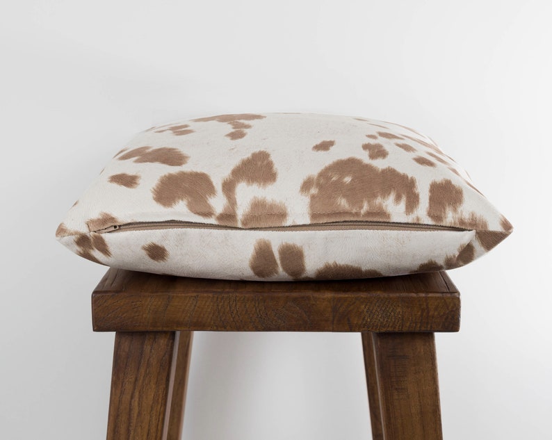 May include: A brown and white cowhide patterned throw pillow with a zipper closure. The pillow is sitting on a wooden stool.