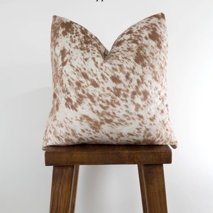 May include: A brown and white cowhide pillow with a salt and pepper pattern. The pillow is sitting on a wooden stool.