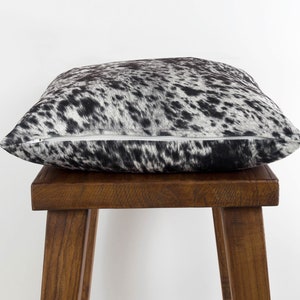 May include: A black and white cowhide pillow with a zipper closure. The pillow is sitting on a wooden stool.