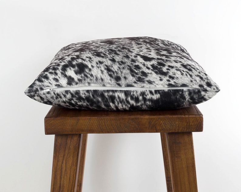 May include: A black and white cowhide pillow with a zipper closure. The pillow is sitting on a wooden stool.