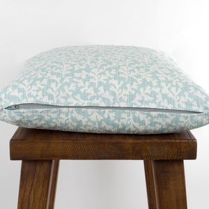 May include: A light blue throw pillow with a white floral pattern. The pillow has a zipper closure and is sitting on a wooden stool.