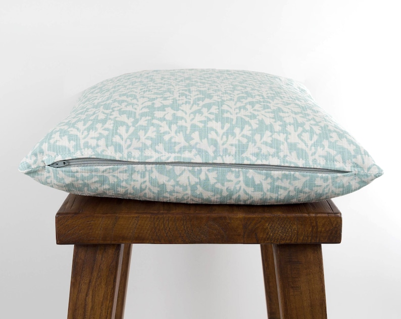 May include: A light blue throw pillow with a white floral pattern. The pillow has a zipper closure and is sitting on a wooden stool.