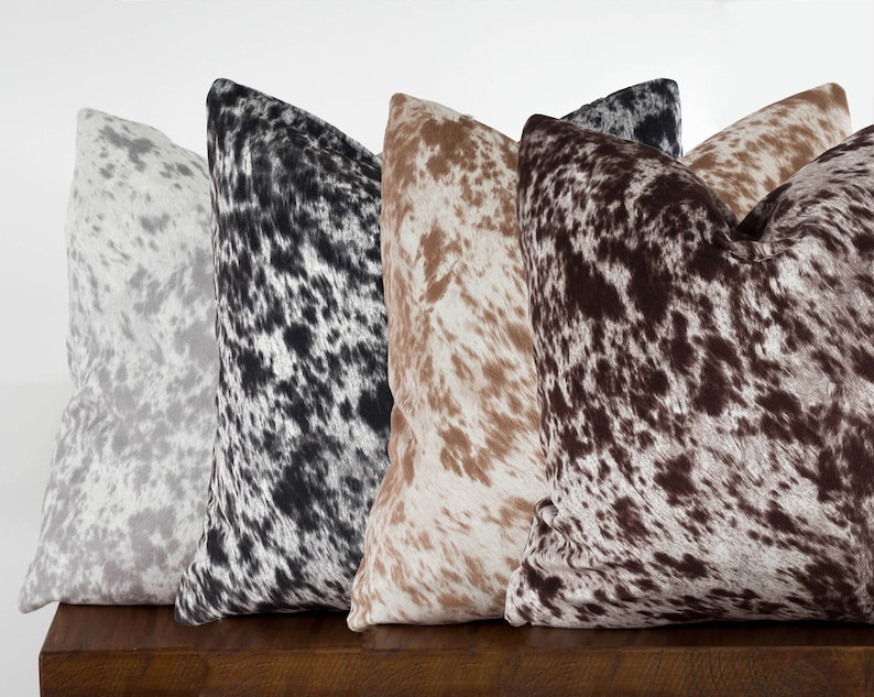 May include: Four decorative throw pillows with a cowhide pattern in different colors: gray, black, brown, and tan. The pillows are arranged in a row on a wooden surface.